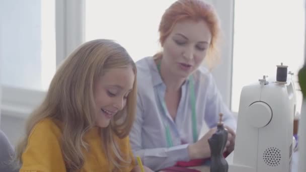 Small girl drawing sitting at the table in the foreground while her red-haired mother sews clothes near in the background. Seamstress works with her daughter. Happy family together. — Stock Video