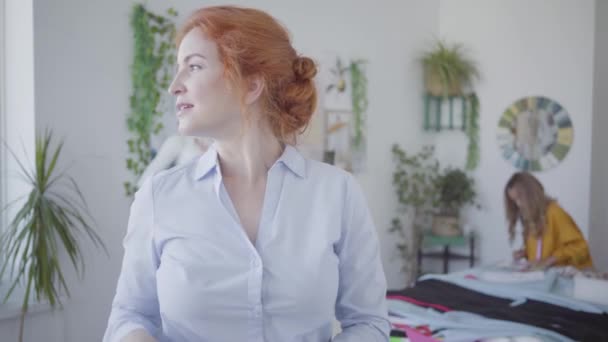 Portrait of attractive red-haired seamstress with the meter on her neck looking in the camera smiling while her little daughter drawing in the background. Happy family working together — Stock Video