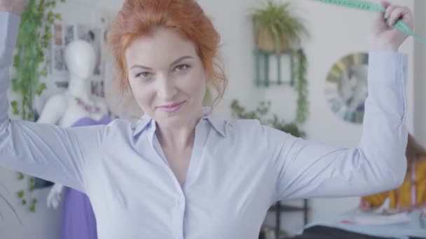 Portrait of attractive red-haired seamstress looking at camera smiling while her little daughter drawing in the background. Happy family working together. — Stock Video