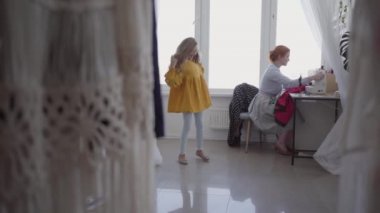 Kızıl saçlı kadın ışık odasında pencerenin yanındaki masada oturan giysilerdiker. Küçük kız yakınlarda dönüyor. Terzi evde çalışıyor. Hobi. Dikiş kıyafetleri