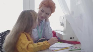 Sevimli küçük kız dikiş dikmeyi öğreniyor. Kızıl saçlı anne kıza elbise için düğmeleri nasıl seçeceğini gösteriyor. Terzi kızıyla evde çalışıyor. Mutlu aile birlikte