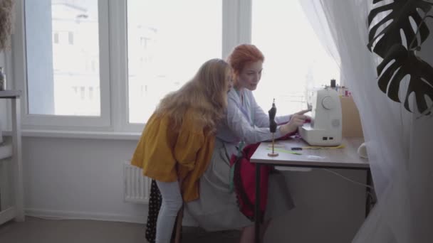 Bela mãe ruiva costura roupas sentadas na mesa perto da janela mostrando sua filha como fazer a costura certa. O Seamstress trabalha em casa. Hobby. Roupa de costura — Vídeo de Stock