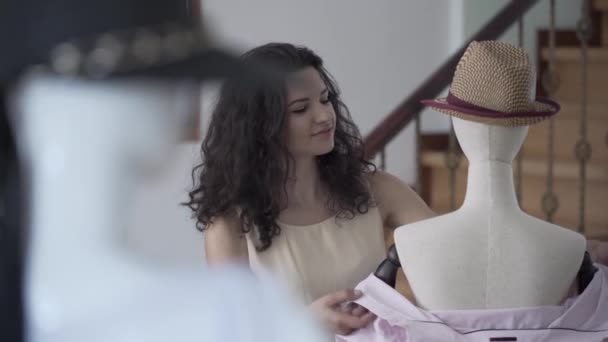 Retrato de jovem sorridente designer de moda feminina ou estilista montagem camisa no manequim com chapéu fedora no showroom ou estúdio — Vídeo de Stock