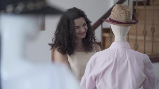 Retrato lindo joven sonriente mujer diseñadora de moda o estilista ajuste camisa en maniquí con sombrero fedora en sala de exposición o estudio — Vídeos de Stock