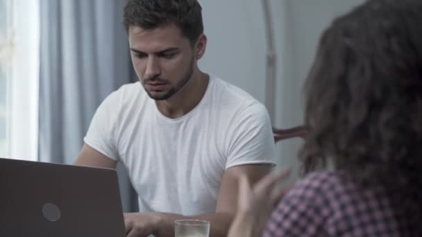 Занятой молодой бородатый мужчина сидит дома за ноутбуком и работает, пока его жена кричит на него. Зависимость от гаджетов. Трудности в отношениях, семейные проблемы — стоковое видео