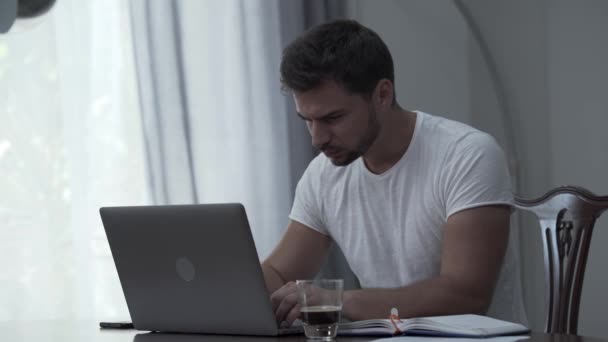 Beyaz tişörtlü sakallı adam evde ya da ofiste kahve içerken dizüstü bilgisayar ve kağıtlarla çalışıyor. Adam yorgun, gözlerini ovuşturuyor ve işini bitiriyor. — Stok video