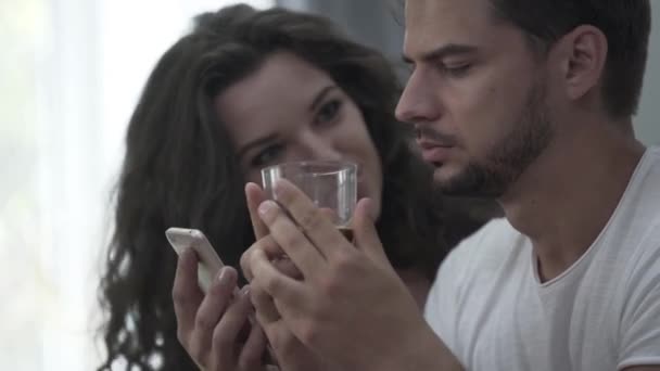 Молодая эмоциональная кудрявая девушка показывает что-то по телефону, что она хочет, чтобы ее парень, но он устал и раздражает — стоковое видео