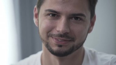 Beyaz tişörtlü yakışıklı gülümseyen adamın portresi kameraya bakarken evde gülümseyerek oturuyor. İçeride dinleniyorum. Işık odasında kendine güvenen sakallı adam