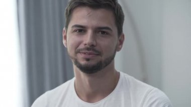 Beyaz tişörtlü gülümseyen adamın kameraya bakarken evde gülümseyerek oturduğu portre. İçeride dinleniyorum. Işık odasında kendine güvenen sakallı adam