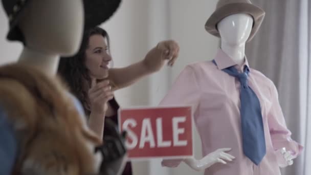 Hübsches Mädchen, das mit dem roten Schild tanzt, auf dem der Verkauf im exklusiven Bekleidungsgeschäft steht. die Frau in der Nähe der modernen Schaufensterpuppen in eleganter Kleidung — Stockvideo