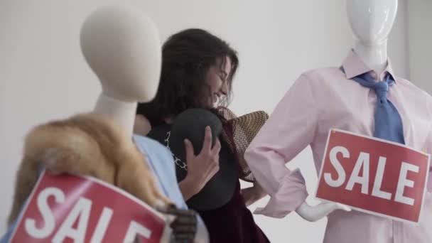 Jeune belle vendeuse bouclée s'amusant dans la boutique ou le showroom avec des mannequins dansant joyeusement avec des chapeaux — Video