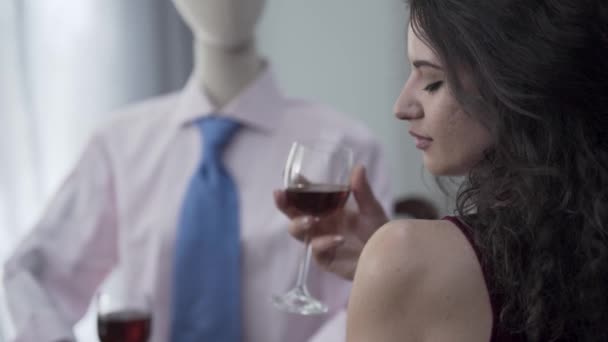 Кудрявая молодая женщина сидит за столом и пьет красное вино с мужским манекеном в шляпе, имитирующим свидание с настоящим мужчиной. Концепция сновидений, воображение, одиночество — стоковое видео