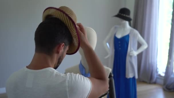 Back view of the young man putting a hat on his head looking at the male mannequin. The guy having fun in the sewing studio or clothes shop — Stock Video
