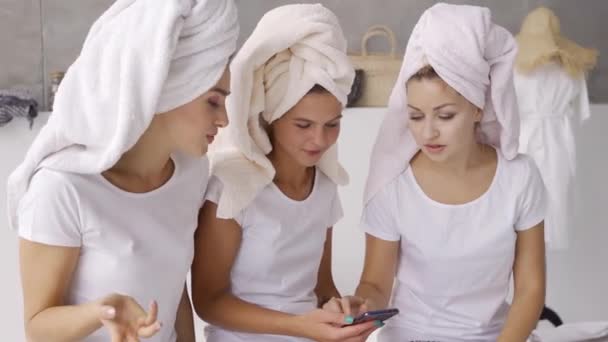 Tres amigas positivas en camisetas blancas con toallas en las cabezas pasando tiempo juntas en la pantalla de desplazamiento del teléfono celular en el baño. Fiesta de gallinas, fiesta de pijamas. Las niñas se divierten en interiores — Vídeo de stock