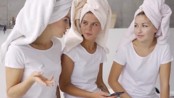 Три позитивні подружки в білих футболках з рушниками на головах спілкуються вдома у ванній кімнаті. Вечірка на піжамі. Дівчата розважаються в приміщенні — стокове відео