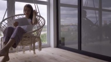 İnce sevimli genç kadın büyük tabandan tavana pencerede onu tablet kullanırken asılı bir sandalyede sallanan. Serbest kavram. Evde dinlenen kadın