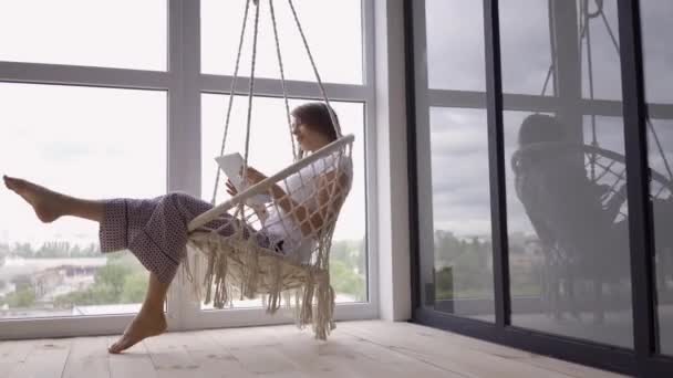 Attraktiv ung kvinna svänger i en hängande stol medan du använder hennes tablett på stora fönster från golv till tak. Frilans koncept. Kvinnan som slappnar av hemma — Stockvideo