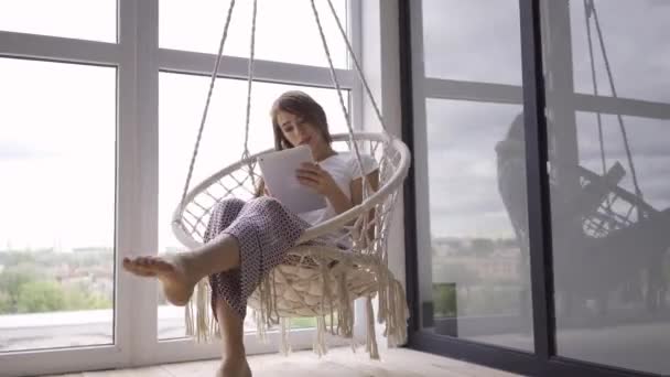 Roztomilá mladá žena houpá se ve visuté židli a používá tabletu ve velkém okně od podlahy ke stropu. Koncept na volné noze. Žena relaxační doma