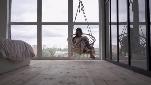 Bedårande ung kvinna svänger i en hängande stol medan du använder hennes tablett på stora fönster från golv till tak. Frilans koncept. Kvinnan som slappnar av hemma — Stockvideo