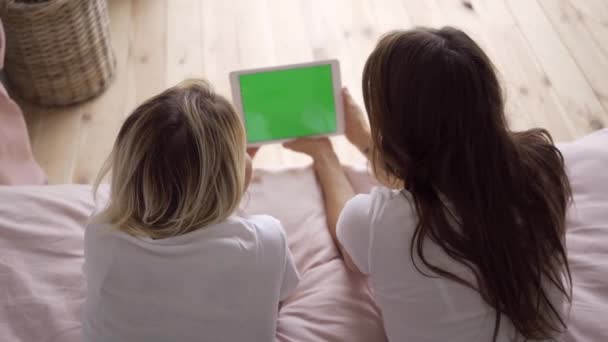 两个穿着白色T恤的年轻女子躺在床上观看平板电脑上的照片的背面。在家休闲。色谱， 绿屏 — 图库视频影像