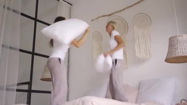 Deux petites amies heureuses dans le même pyjama sautant sur le lit riant et se battant avec des oreillers dans la pièce moderne lumière. Une fête de poules. Loisirs à la maison — Video