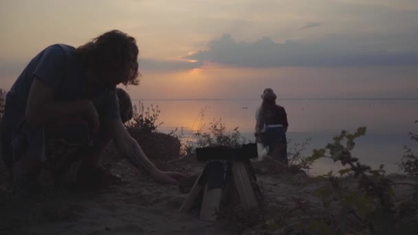 Grupo de jóvenes amigos que se relajan juntos cerca del fuego charlando, abrazando y divirtiéndose con buen humor en el fondo de la impresionante puesta de sol — Vídeos de Stock