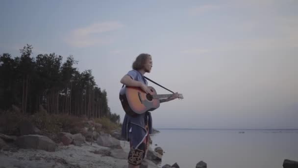 Hipster jonge kerel spelen akoestische gitaar op de achtergrond van avond kleurrijke zonsondergang in de buurt van Summer Beach. — Stockvideo