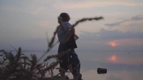 Junger Mann spielt Akustikgitarre vor dem Hintergrund des abends farbenfrohen Sonnenuntergangs in der Nähe des Sommerstrands. — Stockvideo