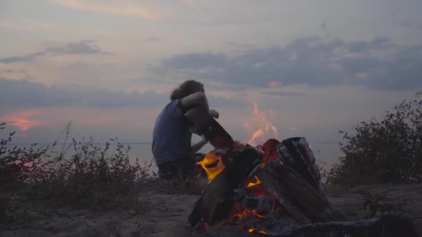 Hipster hombre tocando la guitarra acústica en el fondo de la noche colorido atardecer y playa de verano cerca de hoguera — Vídeo de stock