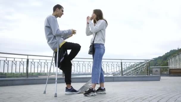 Красивий чоловік з однією ногою на милицях зі скейтбордом розмовляє зі своєю дівчиною на вулиці. Активне життя інвалідів. Хлопець без ноги насолоджується своїм життям. Мотивація, ніколи не здавайся — стокове відео