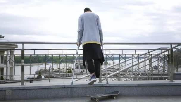 Вид сзади на человека с одной ногой, стоящего на костылях на улице со скейтбордом. Активная жизнь инвалида. Парень без ног наслаждается своей жизнью. Мотивация, никогда не сдаваться — стоковое видео