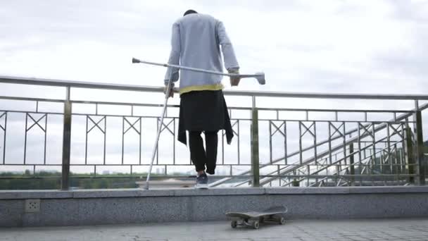 Visão traseira de cara com uma perna de pé em muletas na rua com skate. Vida activa da pessoa com deficiência. O tipo sem pés a gozar a vida. Motivação, nunca desistir — Vídeo de Stock