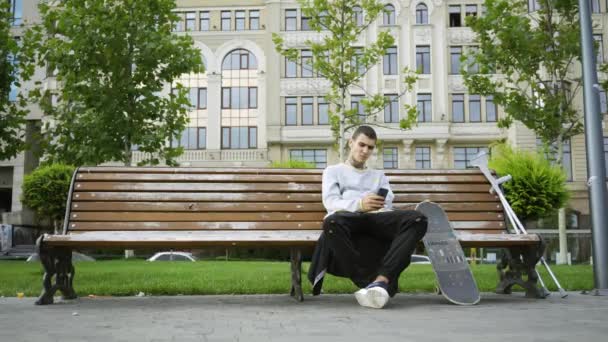 Atrakcyjny człowiek siedzi na ławce w parku słuchania muzyki na jego telefon. W pobliżu znajdują się kule i deskorolka. Aktywne życie osoby niepełnosprawnej. Motywacja, normalne życie, nigdy nie poddawaj się — Wideo stockowe