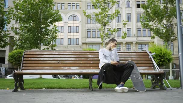 Молодий чоловік сидить на лавці в парку слухаючи музику на своєму мобільному телефоні. Неподалік знаходяться милиці та скейтборд. Активне життя інвалідів. Мотивація, нормальне життя, ніколи не здавайся — стокове відео