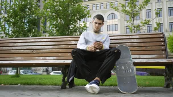 公園のベンチに座って携帯電話で音楽を聴いているハンサムな男。松葉杖とスケートボードが近くにあります。障害者のアクティブな生活。モチベーション、普通の生活、決してあきらめない — ストック動画