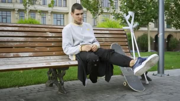 Young man sitting on the bench in the park while listening to music on his cellphone then taking crutches and skateboard and riding away. Active life of disabled person. Motivation, normal life — Stock Video