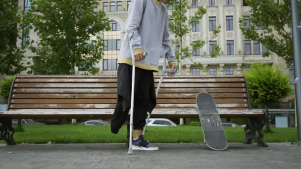 Attraktive Mann sitzt auf der Bank im Park und legt seine Krücken und Skateboard in der Nähe. Das aktive Leben behinderter Menschen. Motivation, normales Leben, niemals aufgeben — Stockvideo