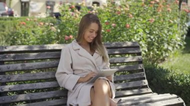 Banyoda sevimli genç kadın parkta bankta oturmuş tablette bilgi kontrolü yapıyor. Güneşli bir günün tadını dışarıda kendine güvenen kız