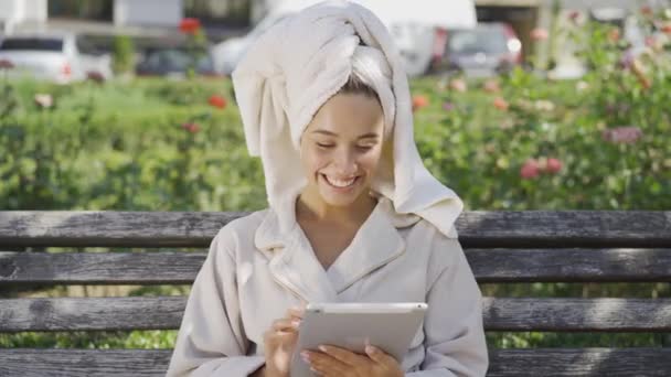Портрет улыбающейся молодой женщины в халате с полотенцем на голове, сидящей на скамейке в парке и проверяющей информацию на планшете. Уверенная девушка наслаждается солнечным днем на открытом воздухе — стоковое видео