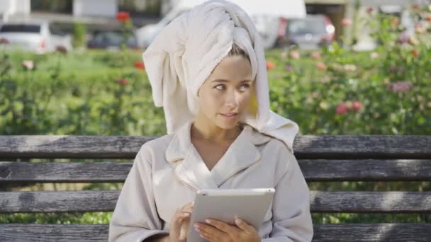 Портрет милой улыбающейся молодой женщины в халате с полотенцем на голове, сидящей на скамейке в парке и проверяющей информацию на планшете. Уверенная девушка наслаждается солнечным днем на открытом воздухе — стоковое видео