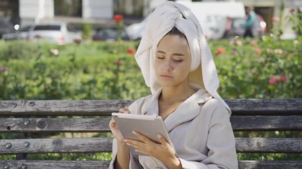 Портрет очаровательной улыбающейся молодой женщины в халате с полотенцем на голове, сидящей на скамейке в парке и проверяющей информацию на планшете. Уверенная девушка наслаждается солнечным днем на открытом воздухе — стоковое видео
