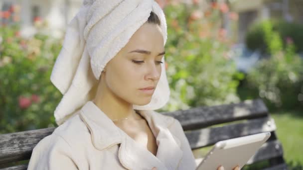 Портрет очаровательной молодой женщины в халате с полотенцем на голове, сидящей на скамейке в парке и проверяющей информацию на планшете. Уверенная девушка наслаждается солнечным днем на открытом воздухе — стоковое видео
