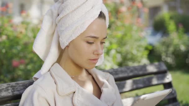 Портрет молодої жінки в халаті з рушником на голові отримав шокуючі новини, сидячи на лавці в парку з планшетом. Впевнена дівчина насолоджується сонячним днем на відкритому повітрі — стокове відео