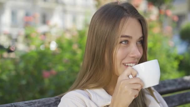 Портрет улыбающейся молодой женщины, сидящей на скамейке в парке и пьющей кофе или чай. Уверенная девушка наслаждается прекрасным утром на открытом воздухе. Беззаботная жизнь — стоковое видео