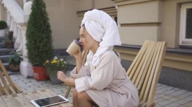 Banyo da sevimli genç kadın başında havlu ile masada kahve içme ve arka bahçesinde tablet bilgi kontrol oturuyor. Güneşli bir günün tadını dışarıda kendine güvenen kız