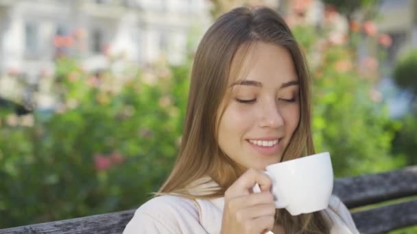 Porträtt av söt leende ung kvinna sitter på bänken i parken dricka kaffe eller te. Säker tjej njuter av vacker morgon utomhus. Bekymmerslöst liv — Stockvideo