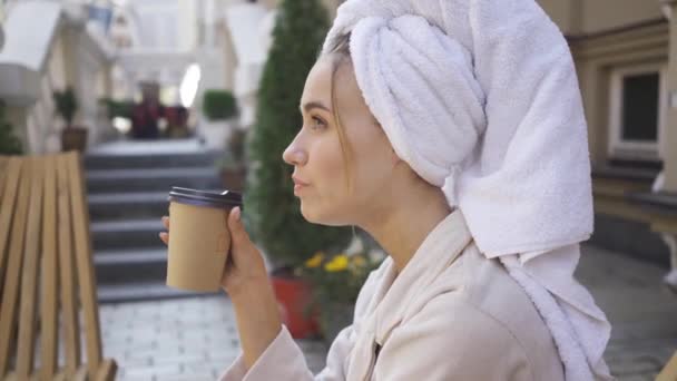 Retrato de mujer joven en albornoz con toalla en la cabeza sentada en la mesa bebiendo café en el patio trasero. Chica rica de confianza disfrutando de un día soleado al aire libre — Vídeo de stock