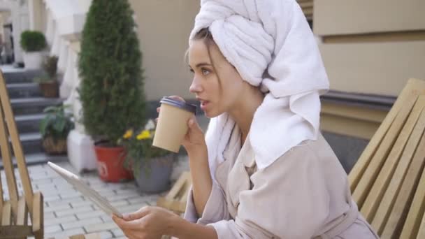バスローブを着た若い女性の肖像画は、コーヒーを飲みながら、裏庭でタブレットを使用してテーブルに座って頭の上にタオルを持っています。屋外で晴れた日を楽しむ自信のある女の子 — ストック動画