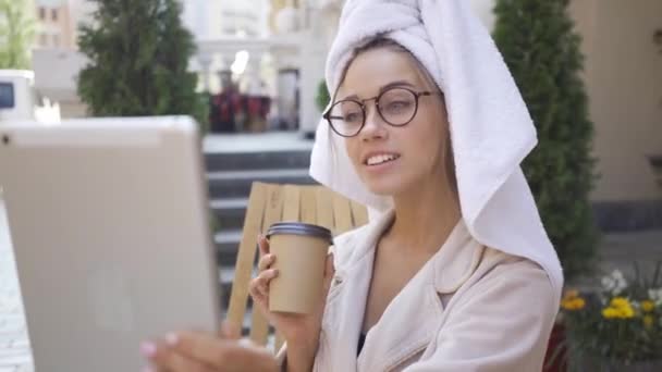 Retrato de mujer joven divertida en gafas y albornoz con toalla en la cabeza mirando la tableta sentada en el patio trasero. Chica segura disfrutando de un día soleado al aire libre . — Vídeos de Stock
