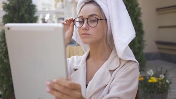 Retrato de la divertida linda mujer joven en gafas y albornoz con toalla en la cabeza mirando la tableta sentada en el patio trasero. Chica segura disfrutando de un día soleado al aire libre . — Vídeos de Stock